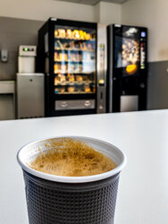 Op de voorgrond een plastic beker met koffie, daarachter een snoep- en koffieautomaat